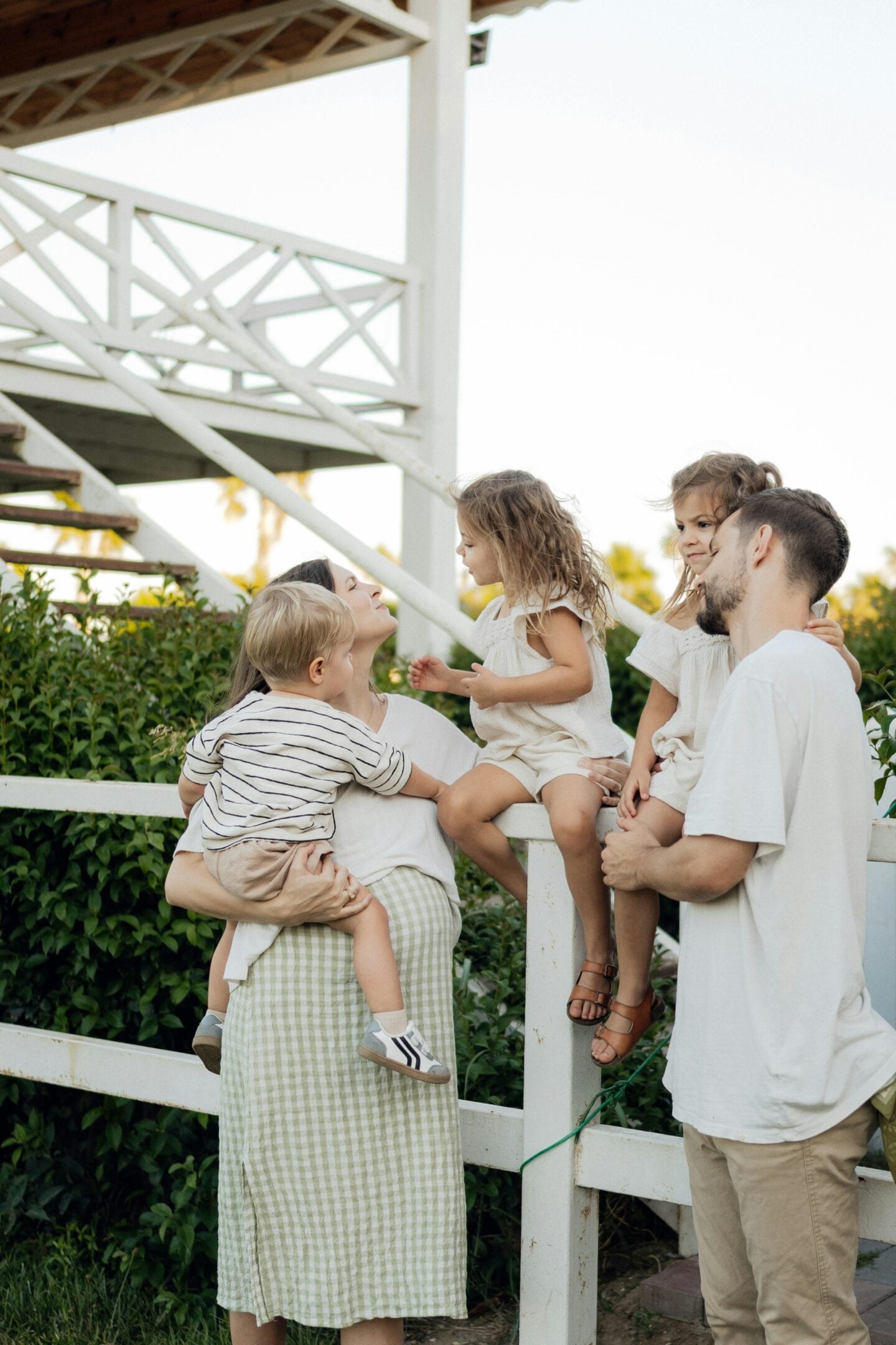 Family of two parents and three young children gathered outdoors by a white wooden fence near a staircase, with the mother holding a toddler while the others sit and stand close together in a relaxed, natural setting.