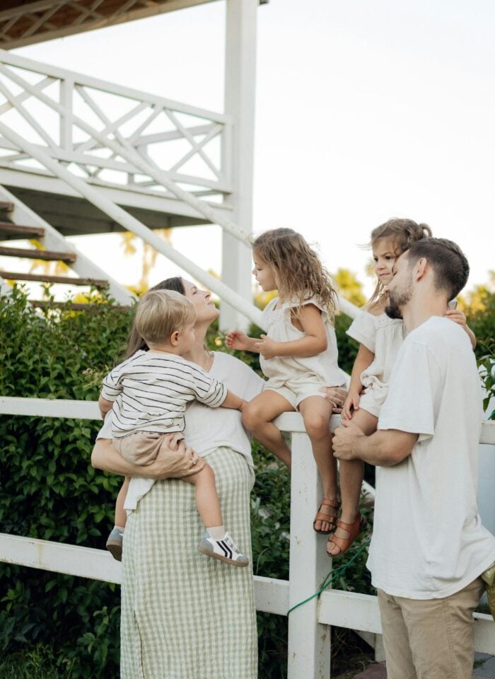 Family of two parents and three young children gathered outdoors by a white wooden fence near a staircase, with the mother holding a toddler while the others sit and stand close together in a relaxed, natural setting.