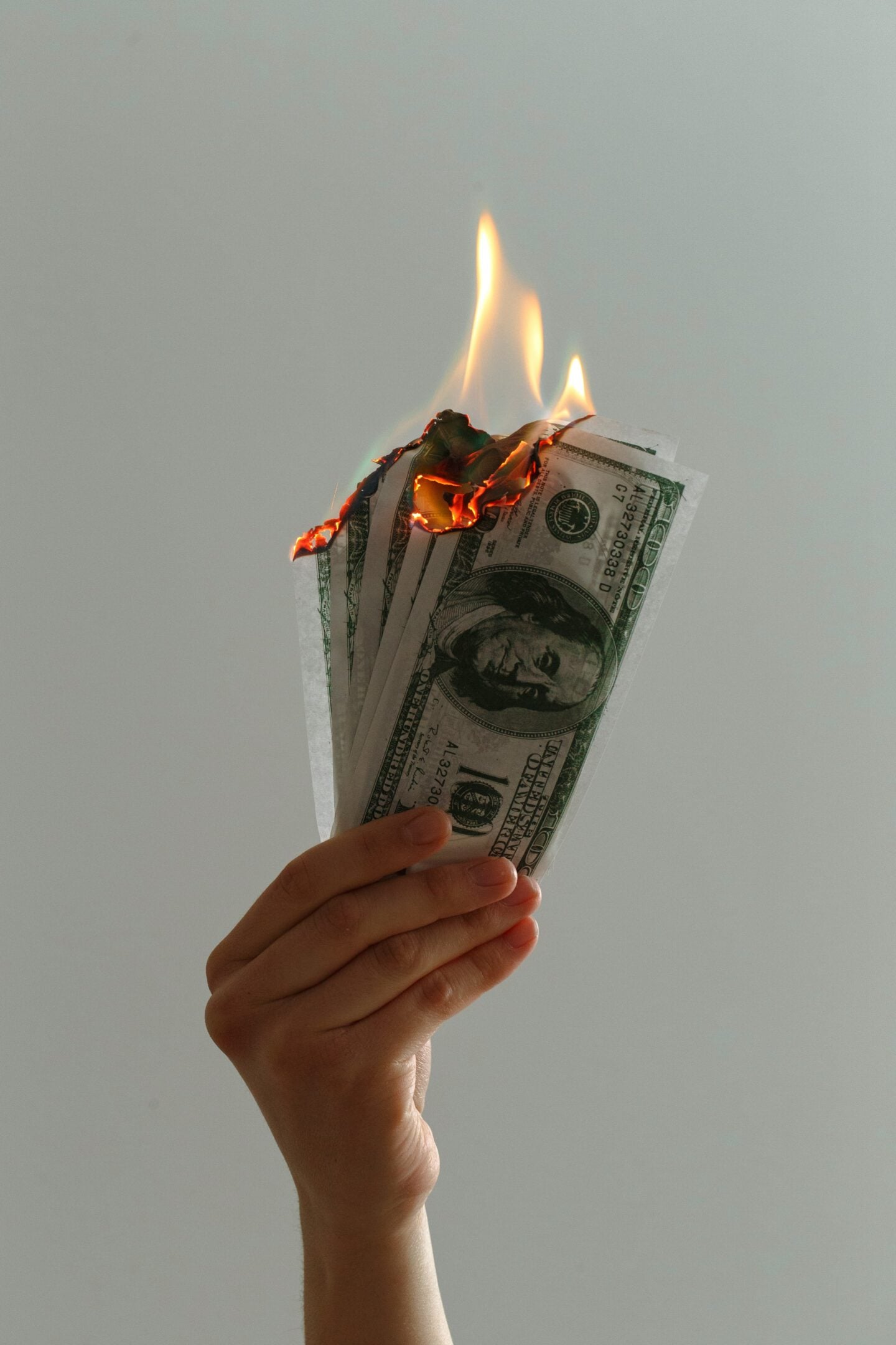 Hand holding several US dollar bills with the top edges on fire, flames burning the money against a plain light background.
