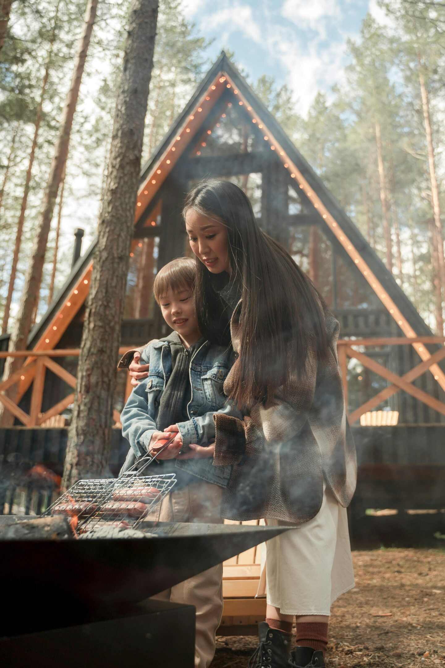 Mother and child grilling food over campfire in forest, cozy family camping moment in front of A-frame cabin, outdoor travel experience with warm lights and nature setting.
