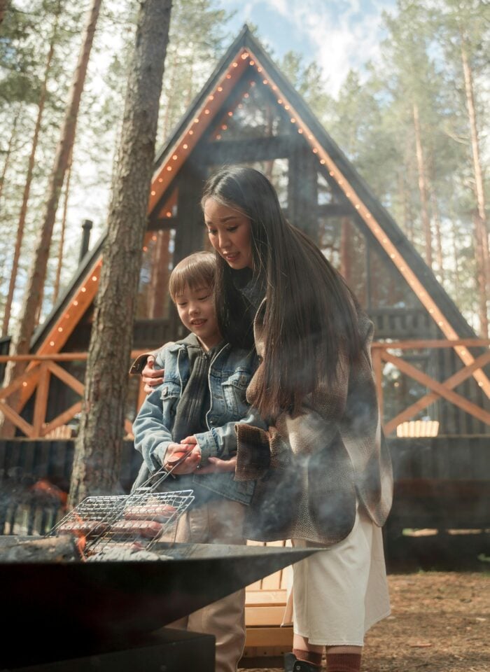 Mother and child grilling food over campfire in forest, cozy family camping moment in front of A-frame cabin, outdoor travel experience with warm lights and nature setting.