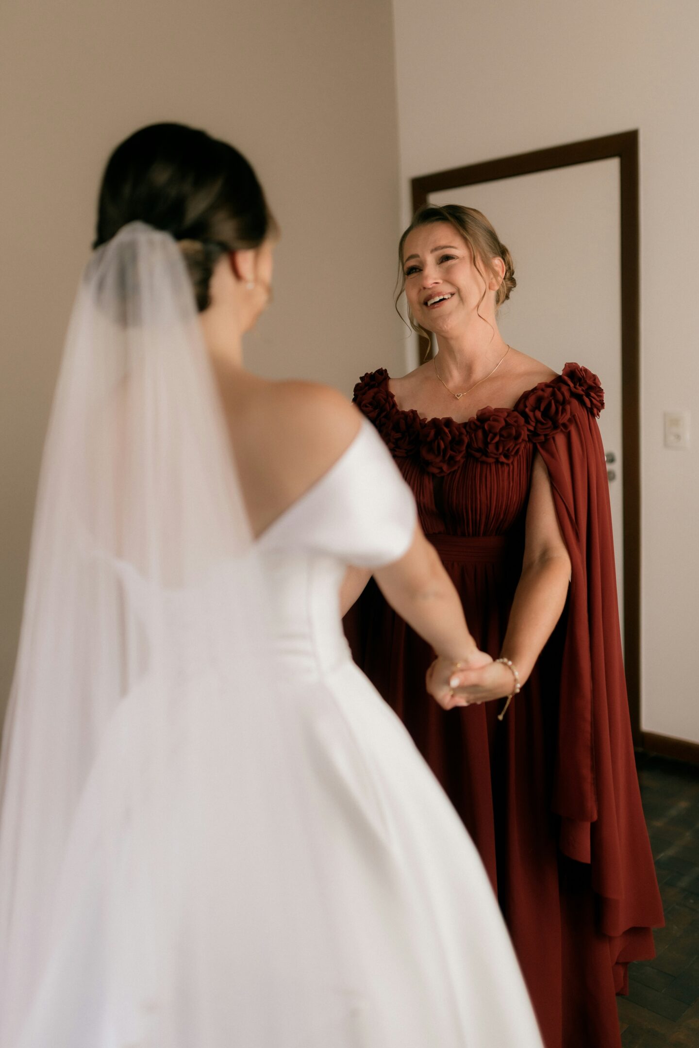 Elegant mother of the bride in a flowing burgundy gown with cape sleeves smiling and holding hands with the bride in a modern white wedding dress and veil, captured in a warm, intimate indoor moment before a desert or resort wedding.