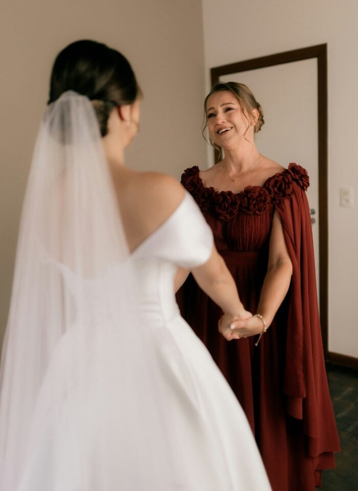 Elegant mother of the bride in a flowing burgundy gown with cape sleeves smiling and holding hands with the bride in a modern white wedding dress and veil, captured in a warm, intimate indoor moment before a desert or resort wedding.