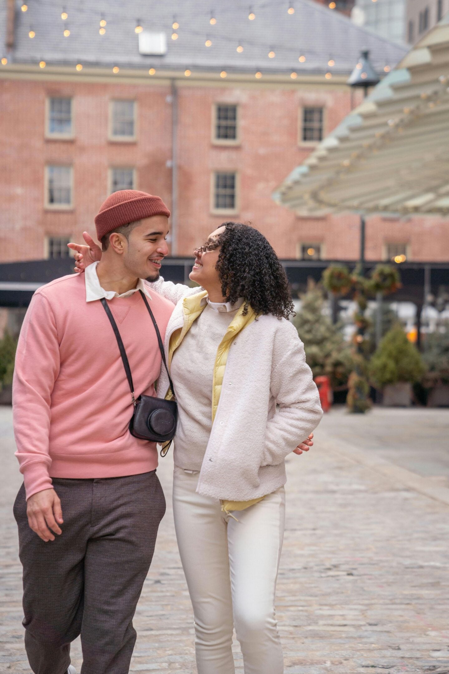 Stylish couple walking arm in arm along a cobblestone street, smiling at each other in a romantic urban setting with warm string lights overhead; the man wears a pink sweater, brown trousers, and a beanie with a camera around his neck, while the woman wears a cream outfit with a light jacket and curly hair, creating a cozy, fashionable street style moment.