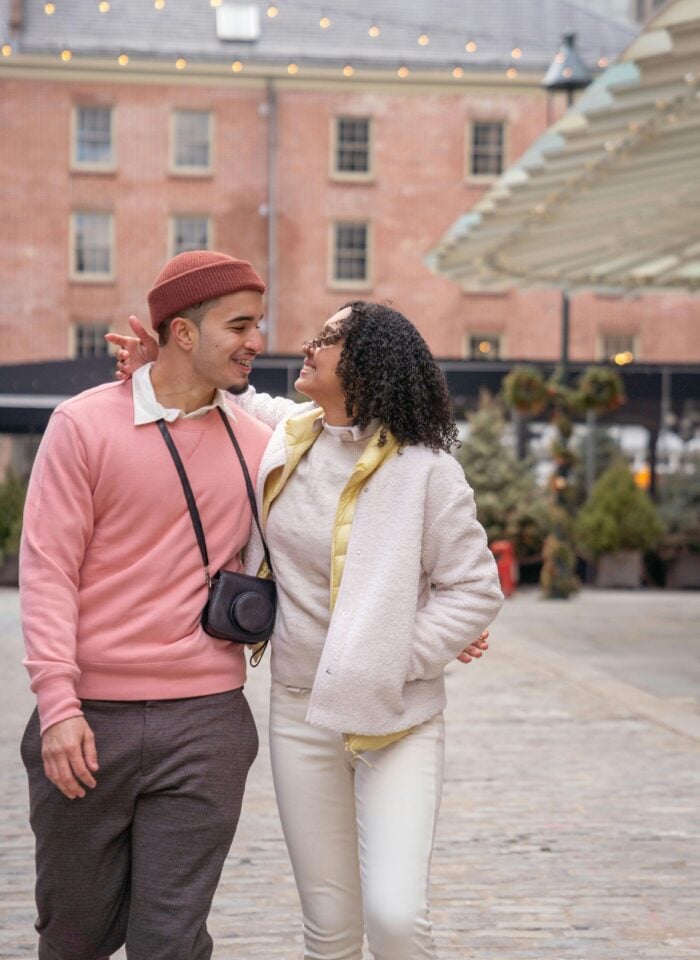 Stylish couple walking arm in arm along a cobblestone street, smiling at each other in a romantic urban setting with warm string lights overhead; the man wears a pink sweater, brown trousers, and a beanie with a camera around his neck, while the woman wears a cream outfit with a light jacket and curly hair, creating a cozy, fashionable street style moment.