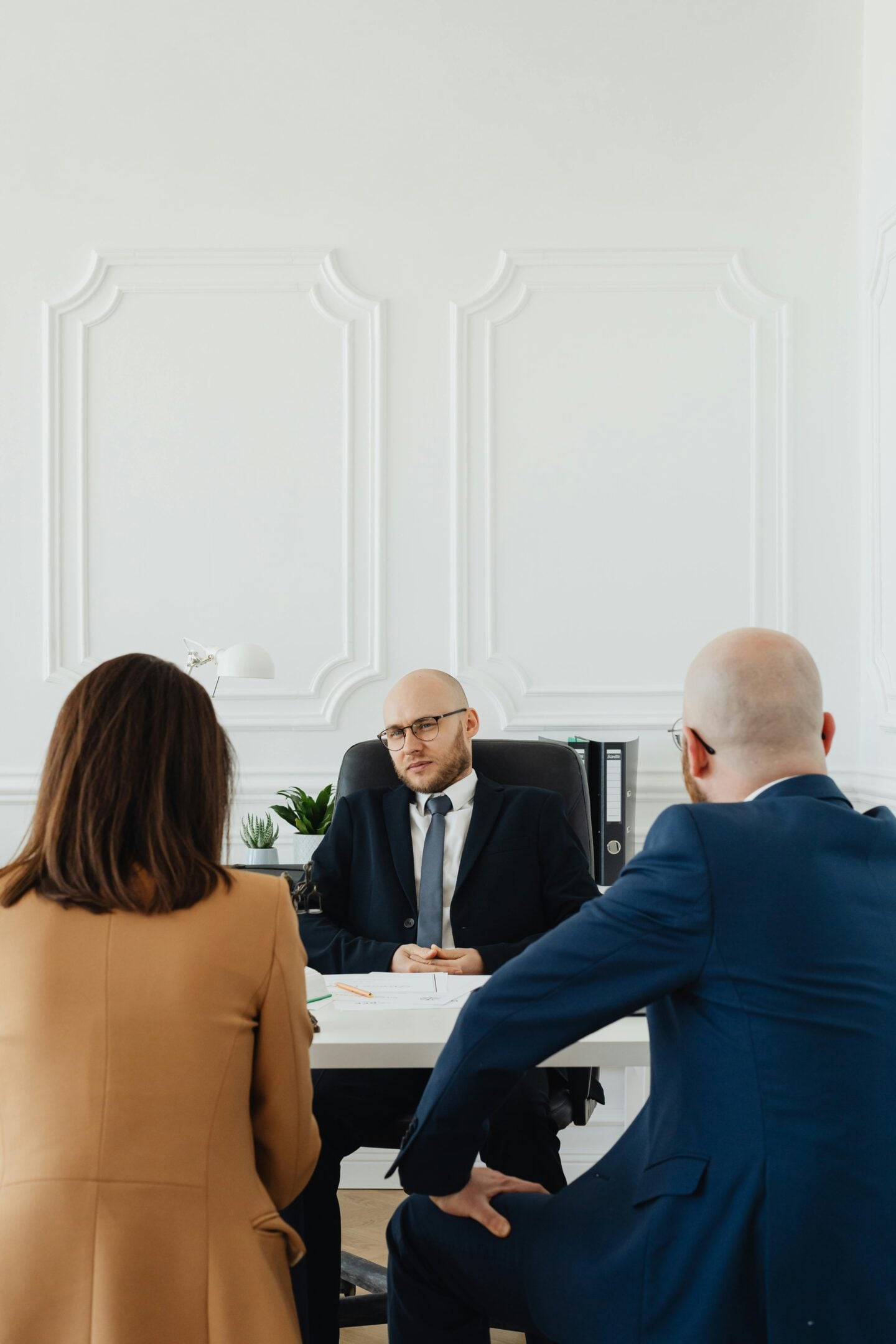 A professionally dressed couple sits across from a lawyer at a desk in a bright, modern office, engaged in a serious consultation with documents placed on the table.