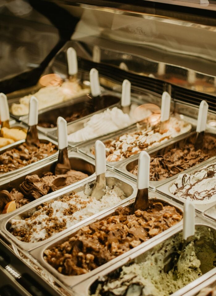 Assorted gelato and ice cream flavors displayed in stainless steel trays with serving scoops in a dessert shop counter.
