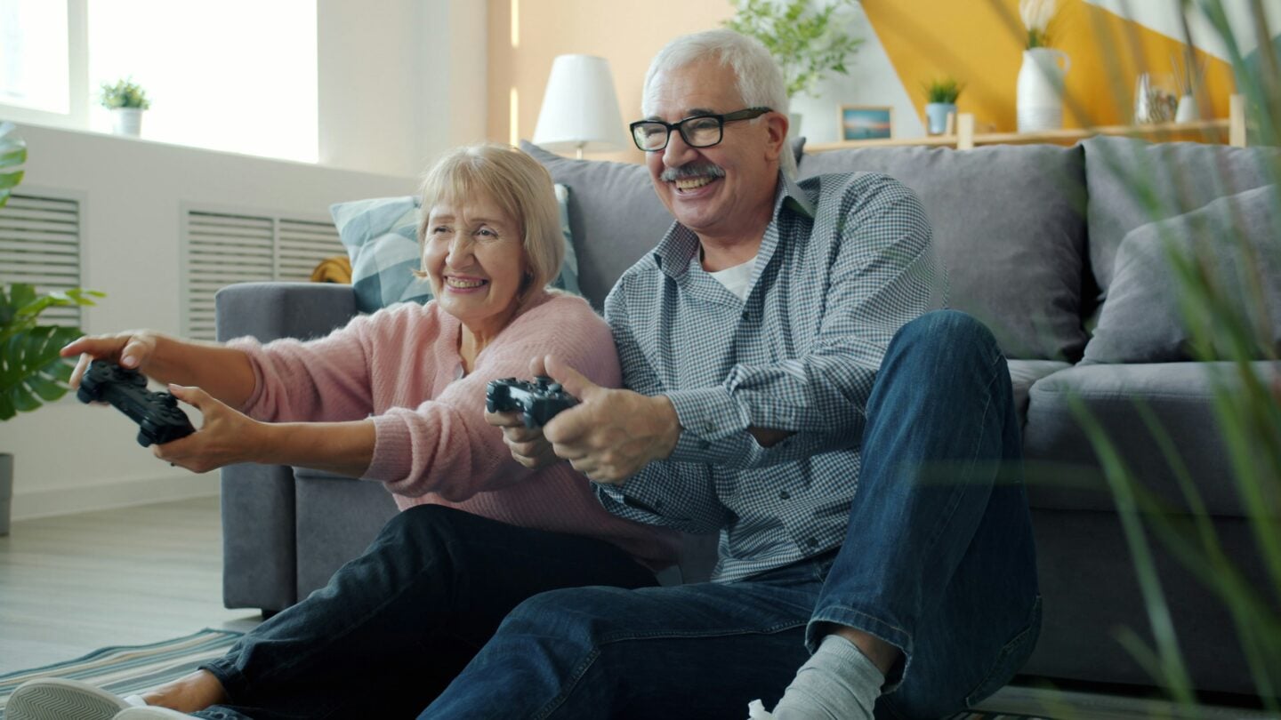 Smiling elderly couple sitting on the floor in a bright modern living room, holding game controllers and playing video games together, enjoying a fun and active moment at home with cozy decor and natural light.