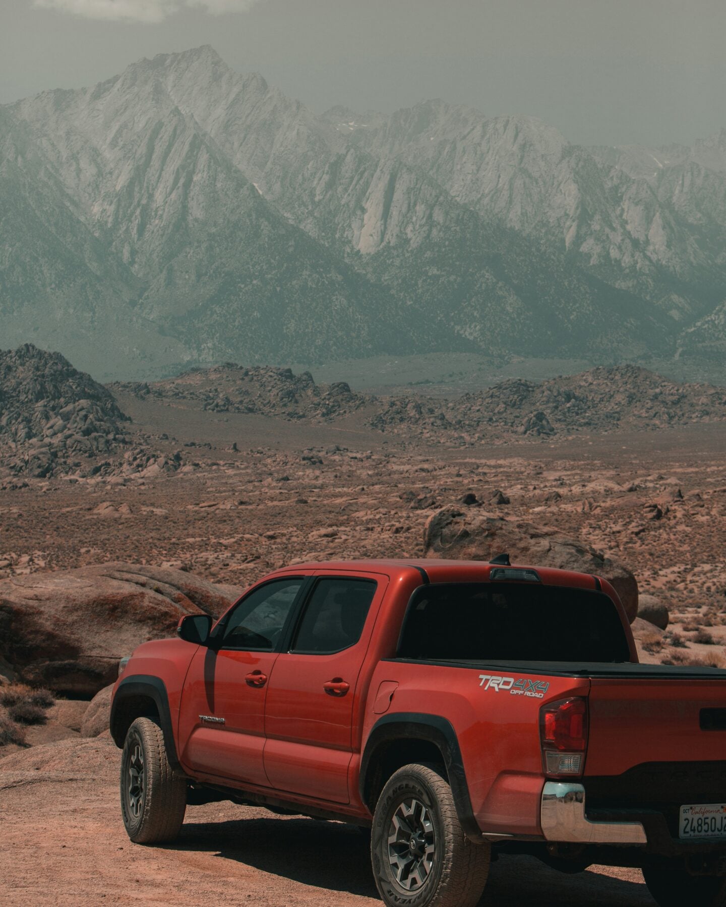 Red pickup truck parked among rocky desert terrain in Alabama Hills, California, with rugged boulder formations in the foreground and dramatic snow-dusted Sierra Nevada mountains rising in the hazy background under a muted sky.