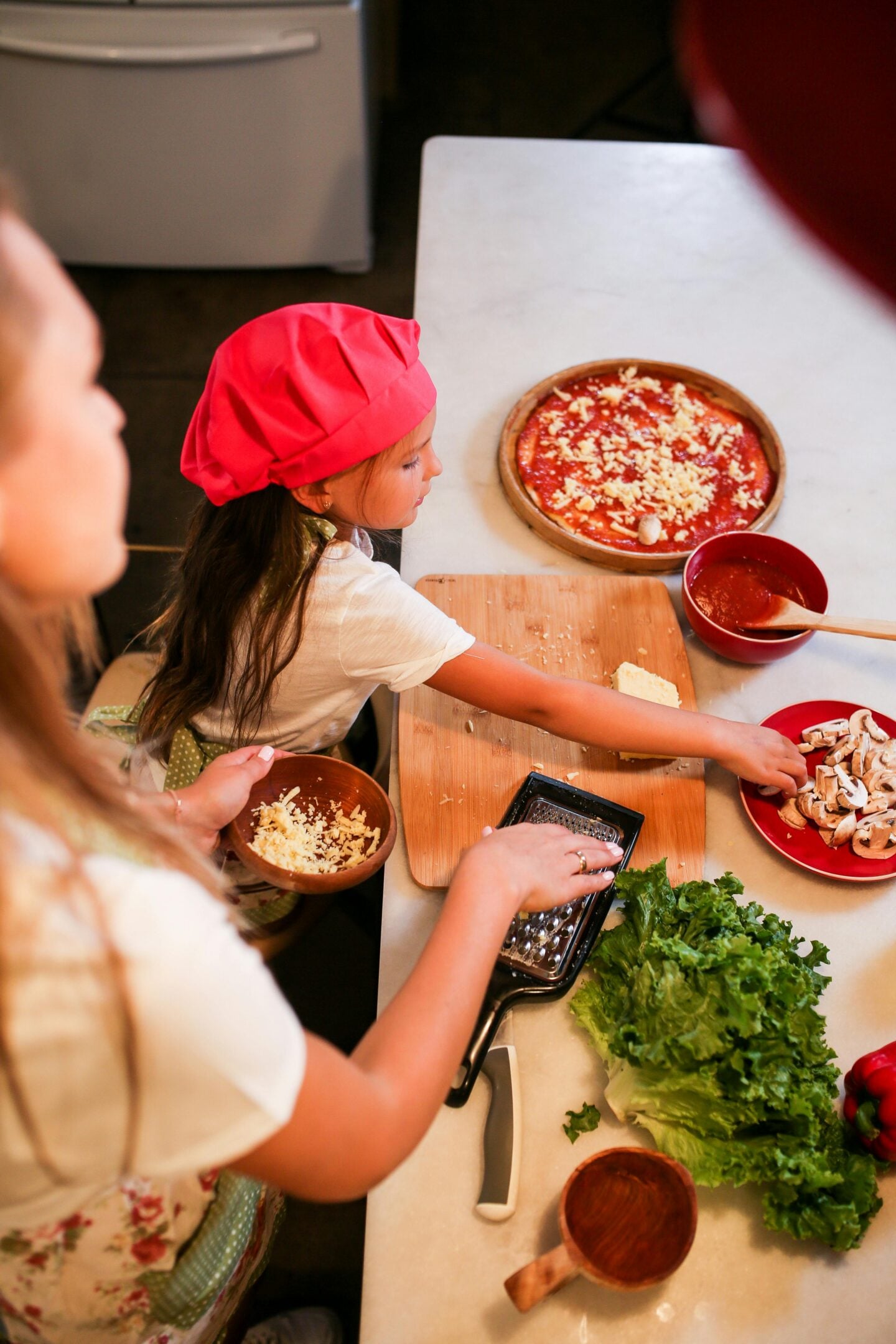 Woman and young girl wearing a red chef hat preparing pizza together in a kitchen, adding toppings like cheese and mushrooms on dough, with fresh vegetables and utensils spread across the counter.