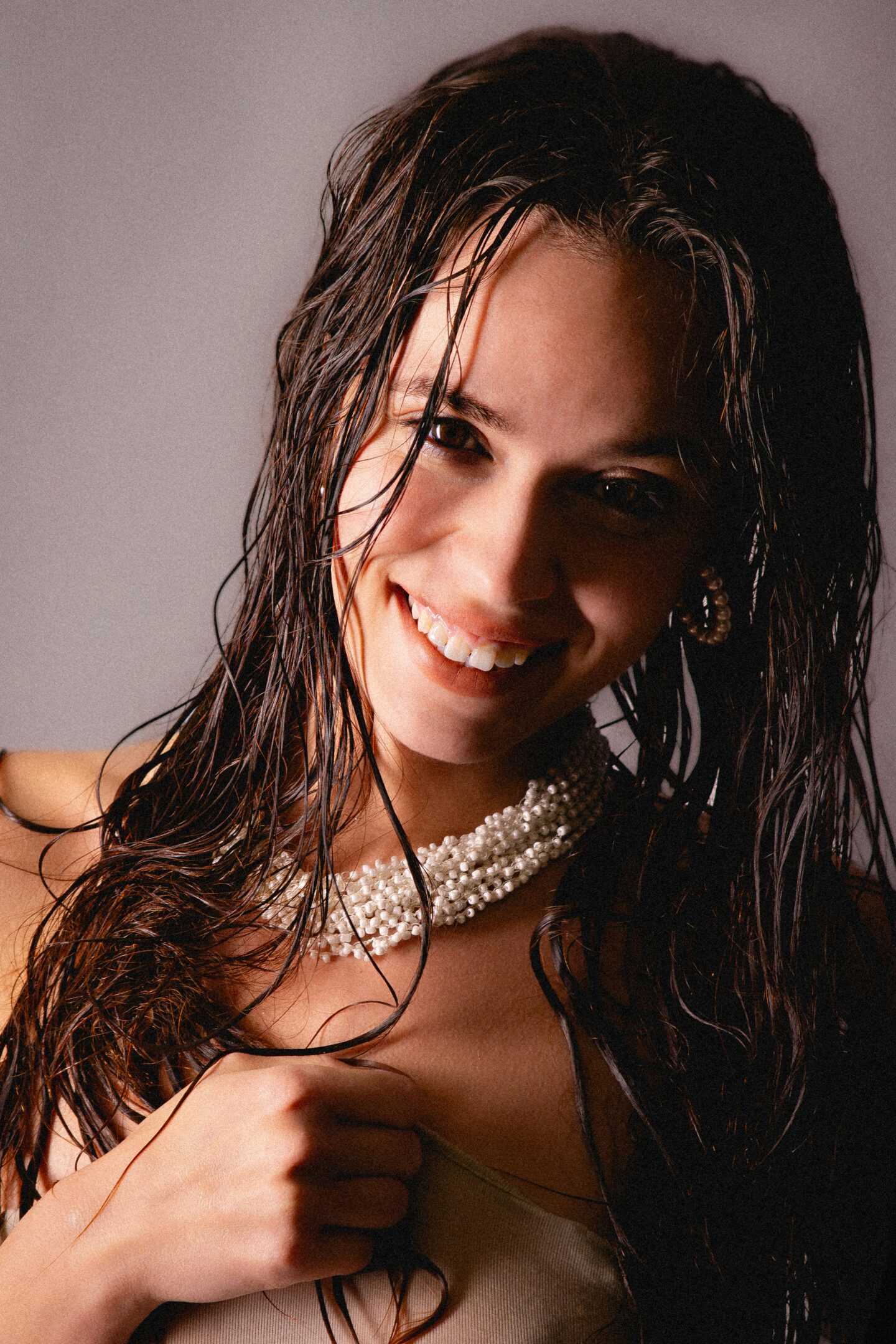 Close-up of a smiling woman with wet hair wearing a pearl necklace and earrings, softly lit against a neutral background.