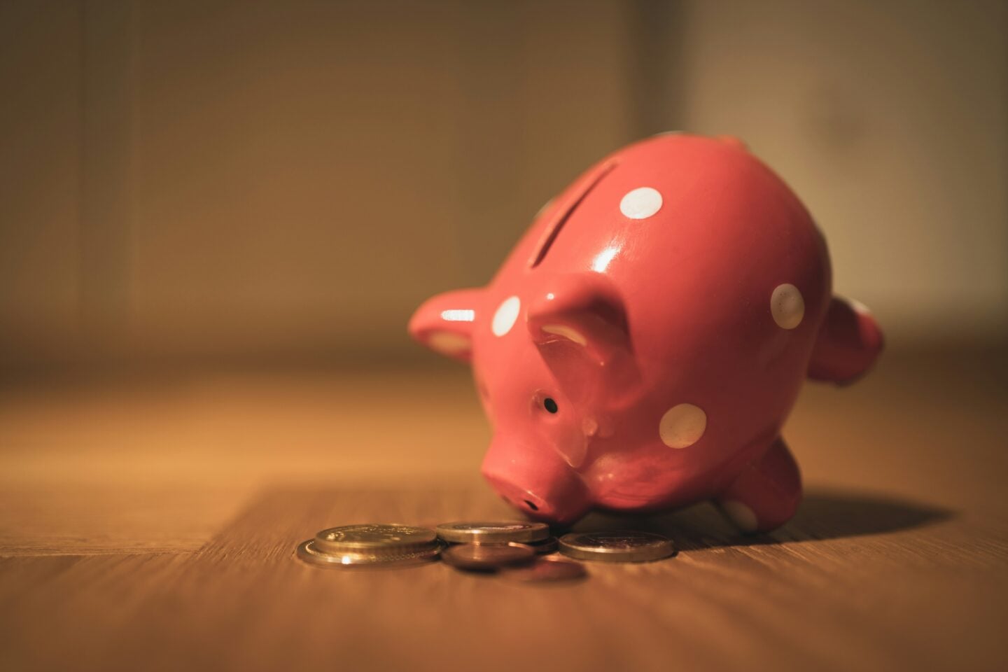 Pink ceramic piggy bank with white polka dots lying on its side on a wooden surface, with several coins scattered in front of it in warm, soft lighting.