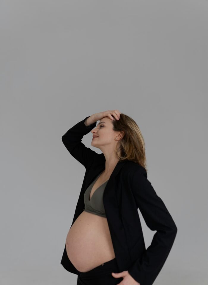 Pregnant woman wearing a black blazer and bra top, gently holding her head and smiling in a minimal studio setting with a neutral background.
