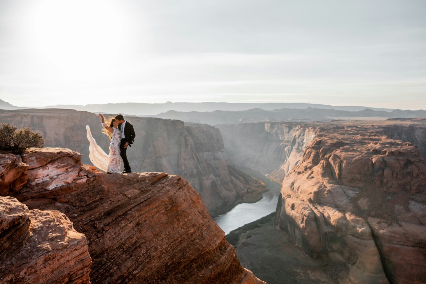 Romantic desert wedding scene with bride and groom standing on a dramatic canyon cliff edge overlooking a winding river, golden hour lighting illuminating red rock landscapes and flowing bridal veil in a breathtaking Arizona elopement setting.