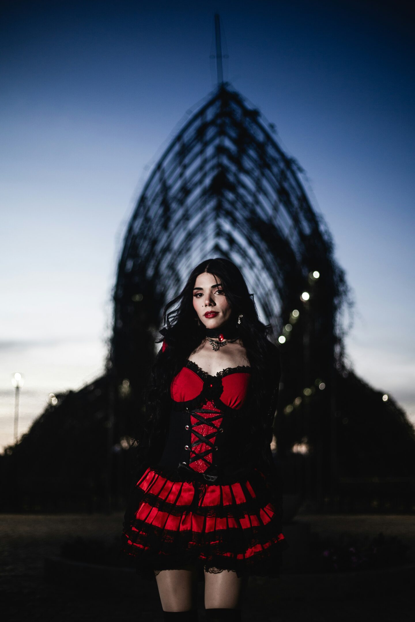 Woman in red and black gothic dress standing outdoors at dusk, dramatic corset outfit with lace details and dark aesthetic, moody evening setting with architectural structure in background.