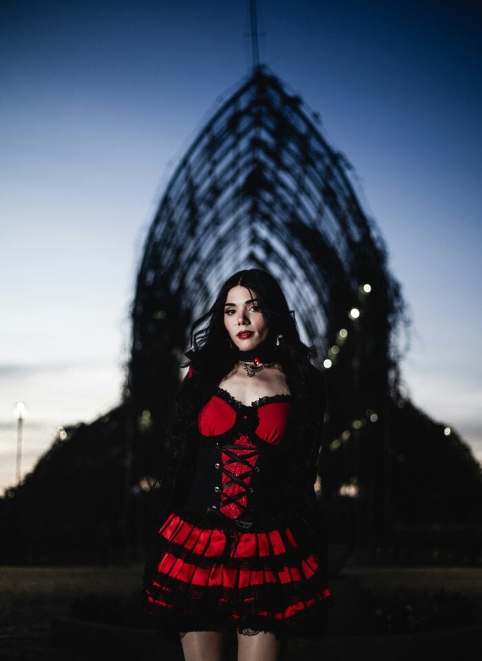 Woman in red and black gothic dress standing outdoors at dusk, dramatic corset outfit with lace details and dark aesthetic, moody evening setting with architectural structure in background.