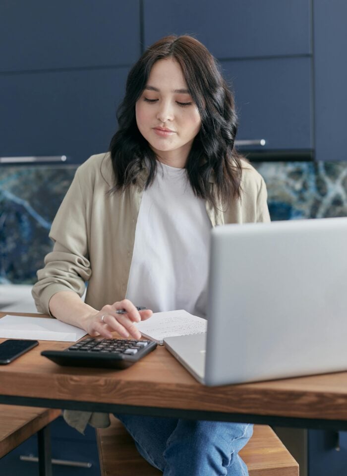 A young woman sitting at a wooden table, using a calculator while working on a laptop, with a notebook and smartphone beside her in a modern kitchen setting.