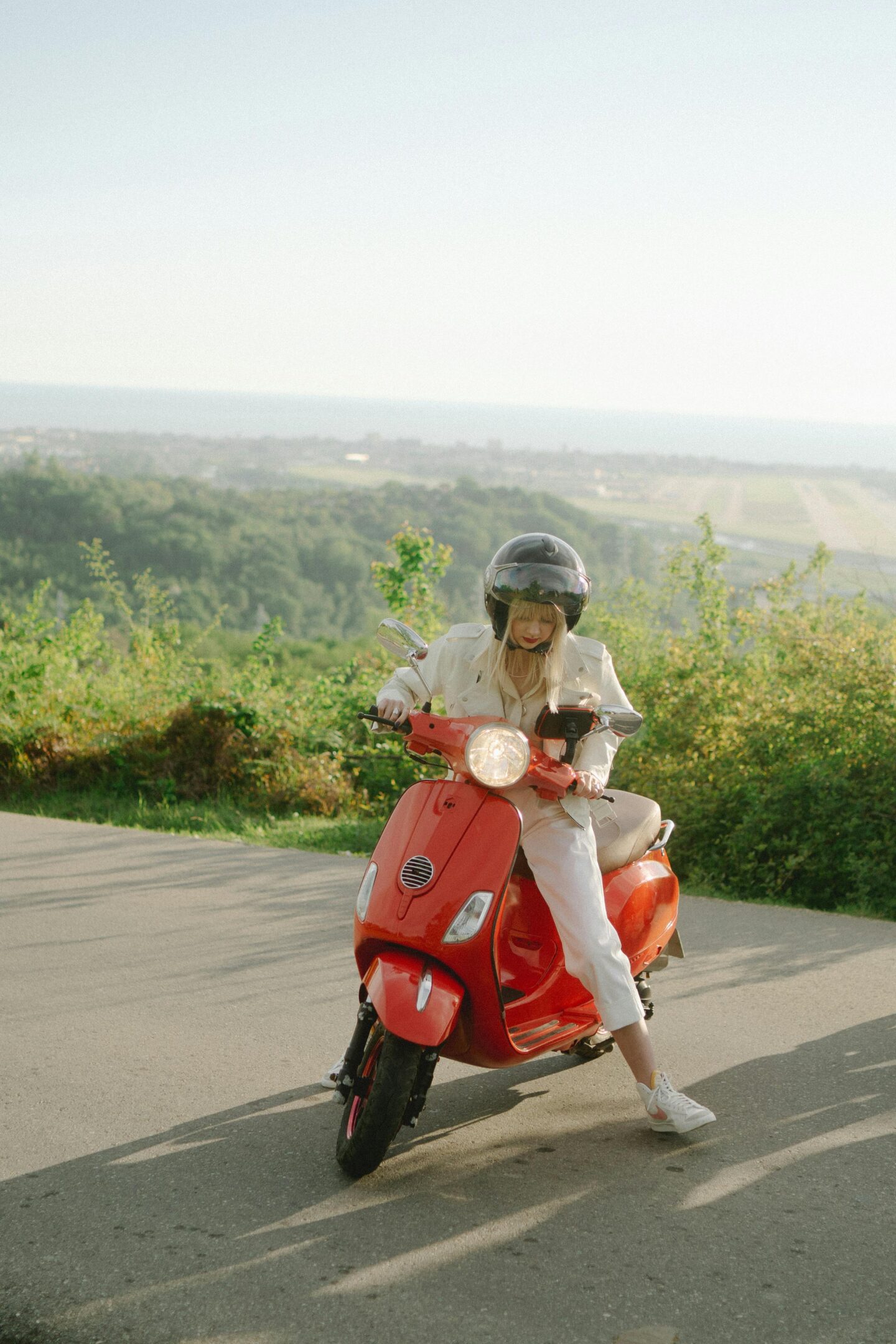 Woman riding a red scooter on a scenic countryside road, wearing helmet and casual outfit, stylish travel moment with sunny landscape and open view in background. 