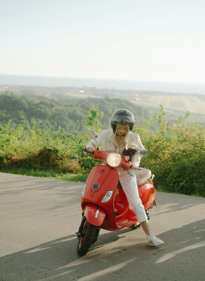 Woman riding a red scooter on a scenic countryside road, wearing helmet and casual outfit, stylish travel moment with sunny landscape and open view in background.