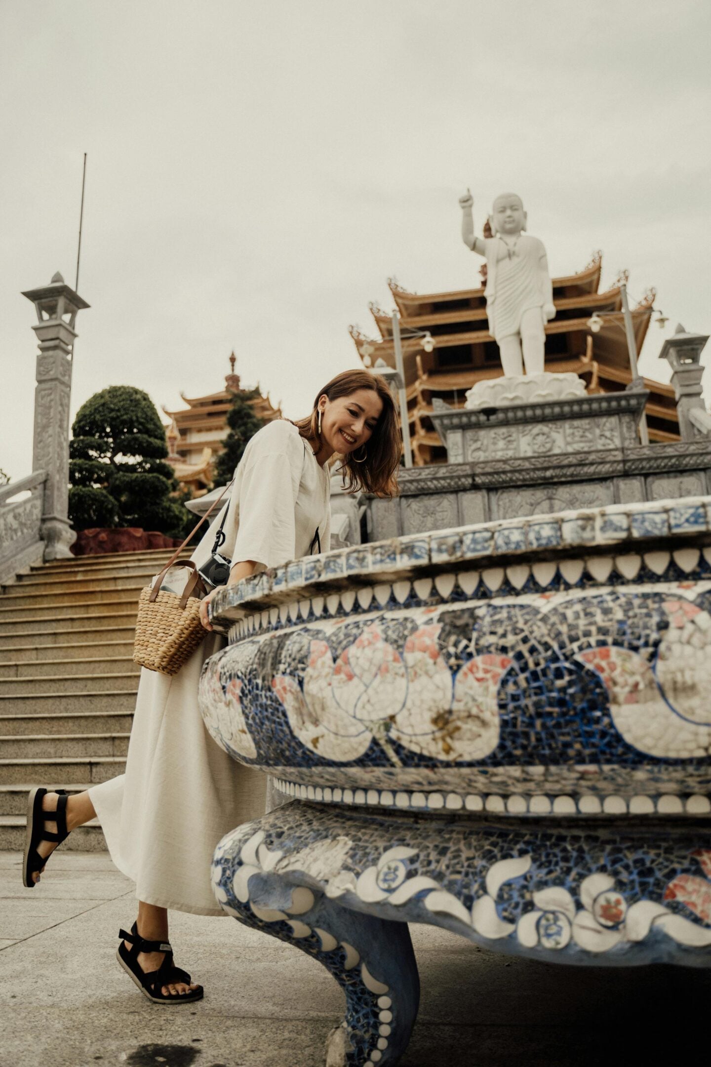 Smiling woman in a light midi dress and black walking sandals leaning over a decorative mosaic basin at an ornate temple, with stone steps, traditional pagoda-style architecture, and a large statue in the background.