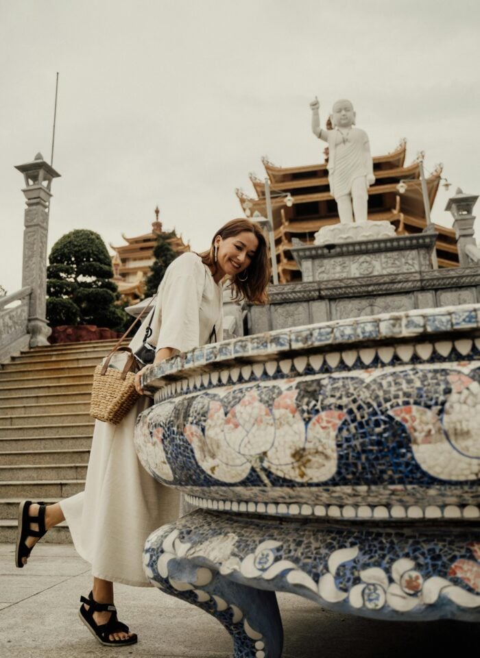 Smiling woman in a light midi dress and black walking sandals leaning over a decorative mosaic basin at an ornate temple, with stone steps, traditional pagoda-style architecture, and a large statue in the background.
