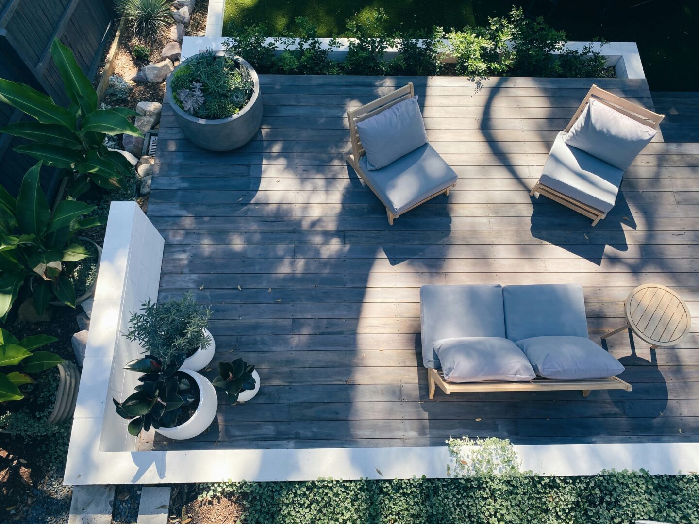Modern backyard deck with cushioned outdoor lounge furniture, potted plants, and minimalist landscaping viewed from above in natural sunlight.