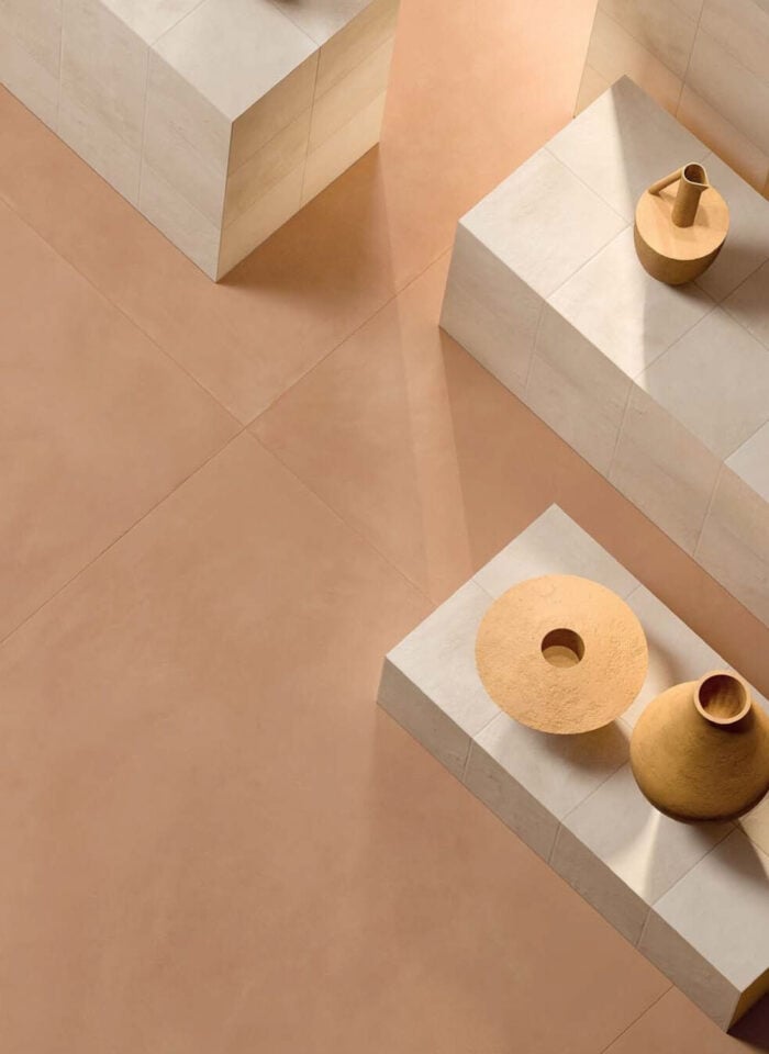 Minimalist interior design featuring warm beige Italian porcelain floor tiles with sculptural ceramic vases displayed on geometric stone pedestals in soft natural light.