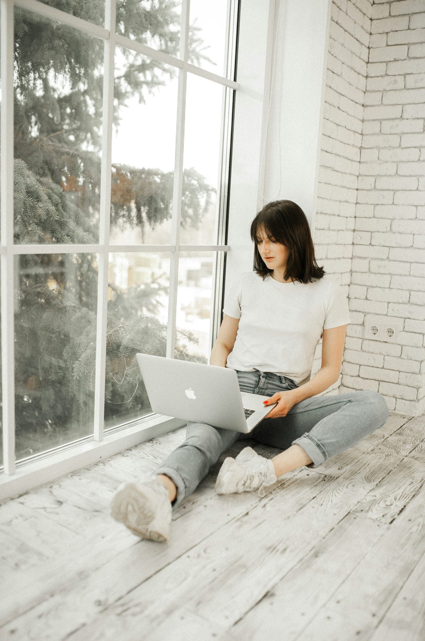 Woman sitting on a wooden floor by a large window using a laptop in a bright minimalist home interior.