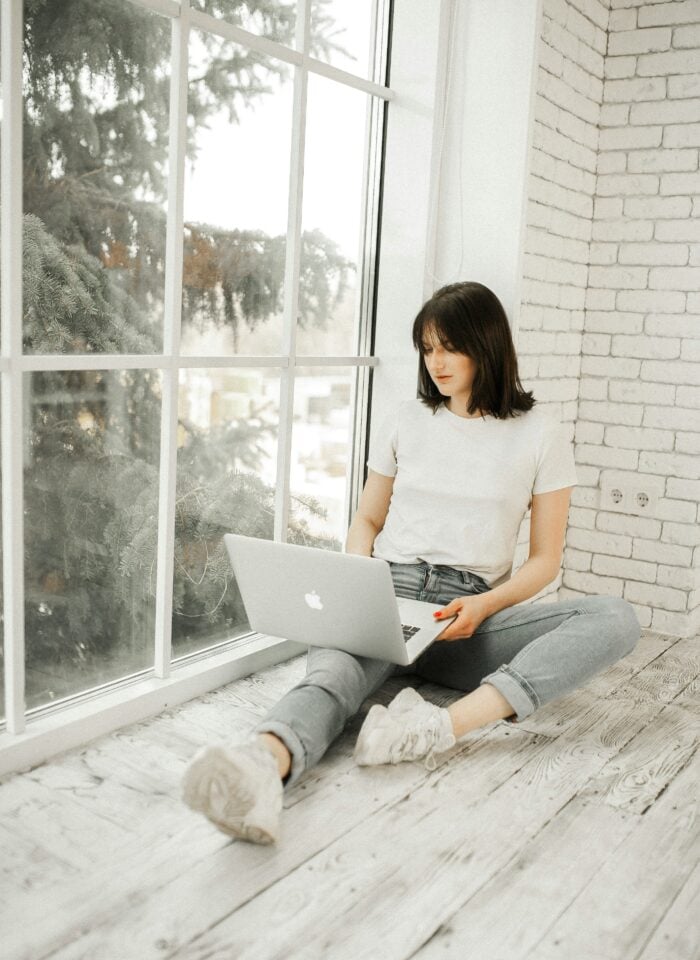 Woman sitting on a wooden floor by a large window using a laptop in a bright minimalist home interior.
