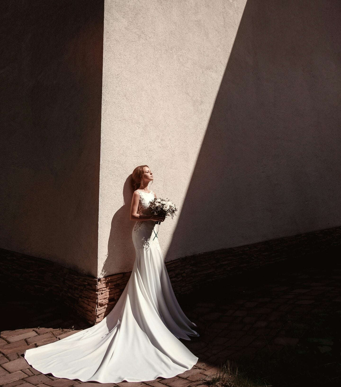 Elegant bride in a fitted white designer wedding gown standing against a dramatic sunlit wall with architectural shadows, holding a bouquet during an editorial-style bridal portrait.