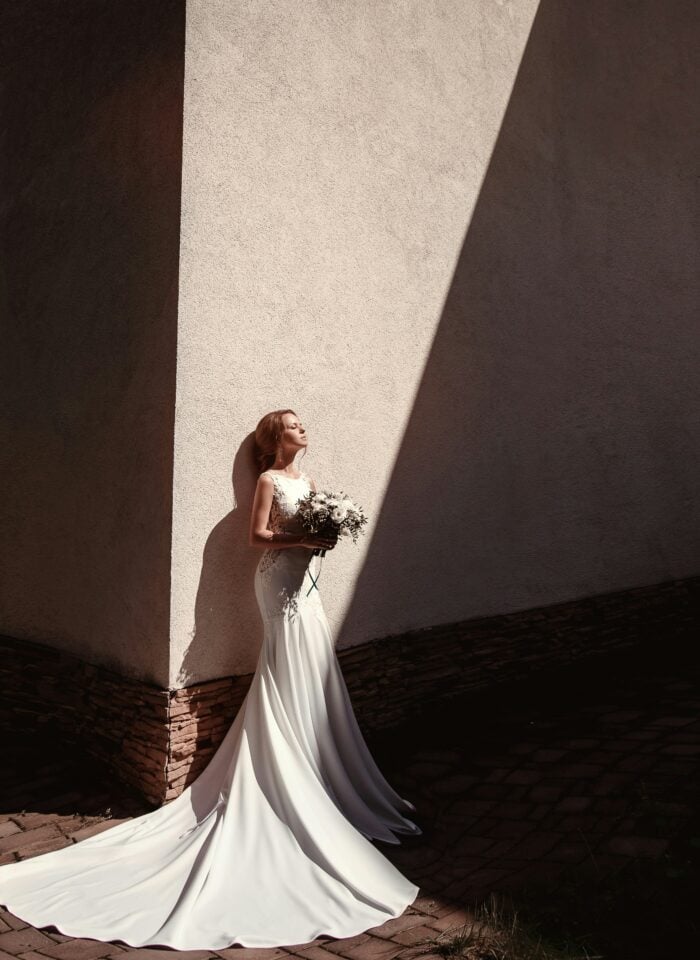 Elegant bride in a fitted white designer wedding gown standing against a dramatic sunlit wall with architectural shadows, holding a bouquet during an editorial-style bridal portrait.
