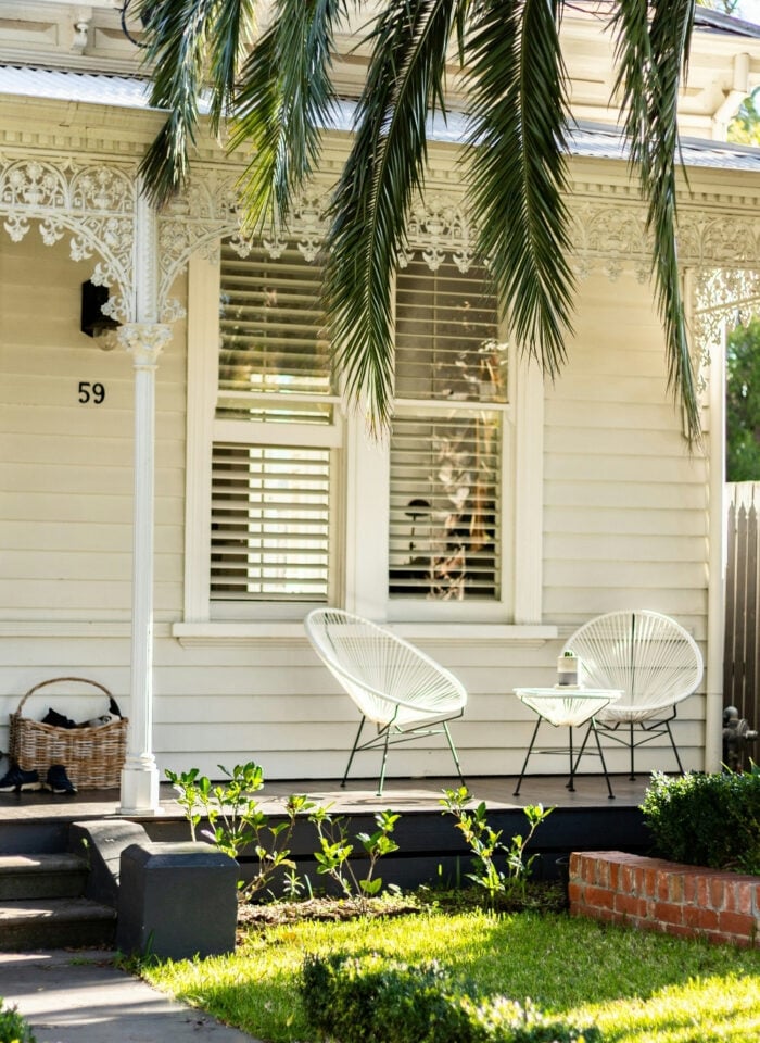 Front view of a white Victorian-style house with decorative trim, palm trees, a manicured lawn, and a shaded porch featuring modern white outdoor chairs.