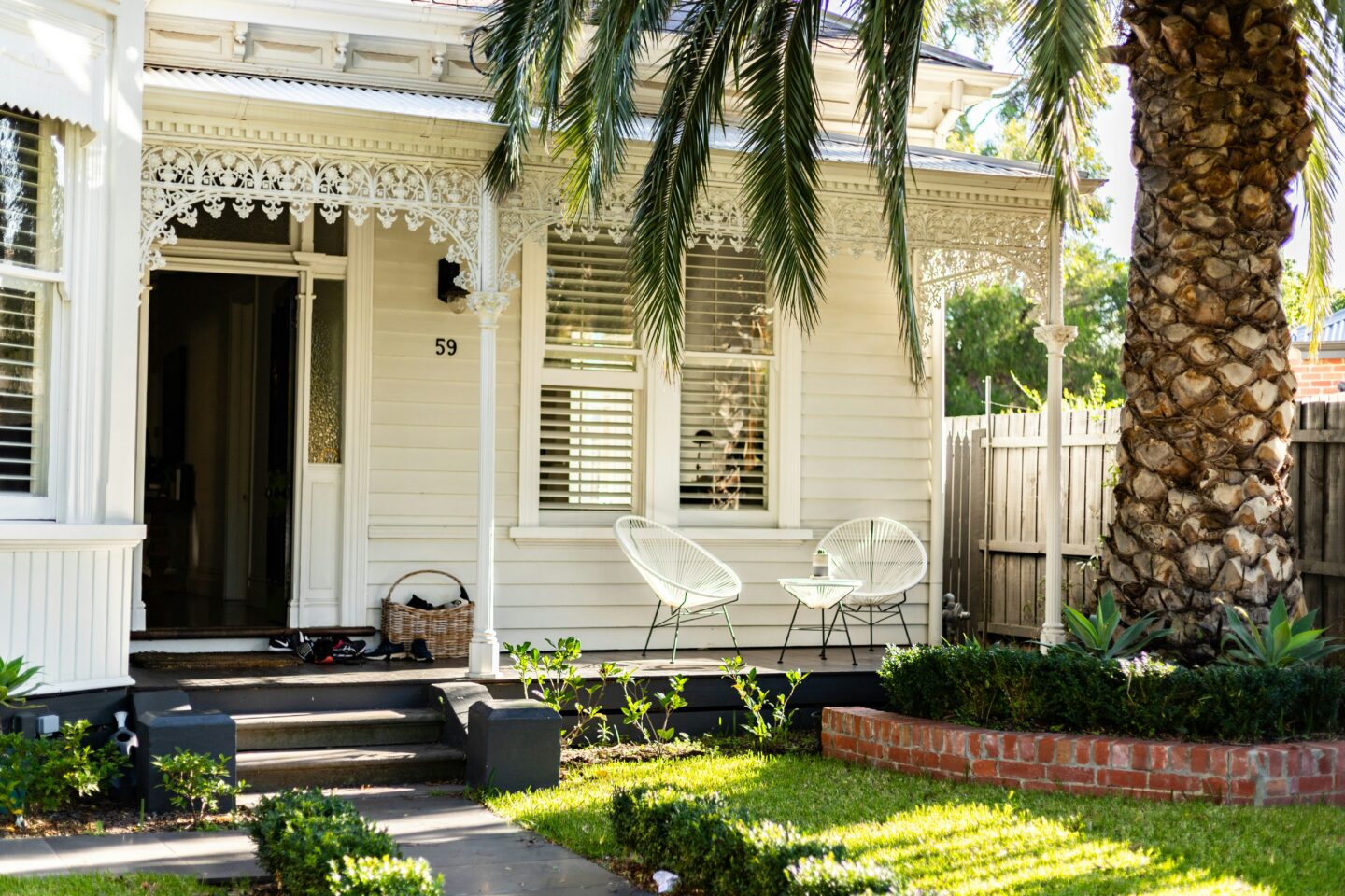 Front view of a white Victorian-style house with decorative trim, palm trees, a manicured lawn, and a shaded porch featuring modern white outdoor chairs.