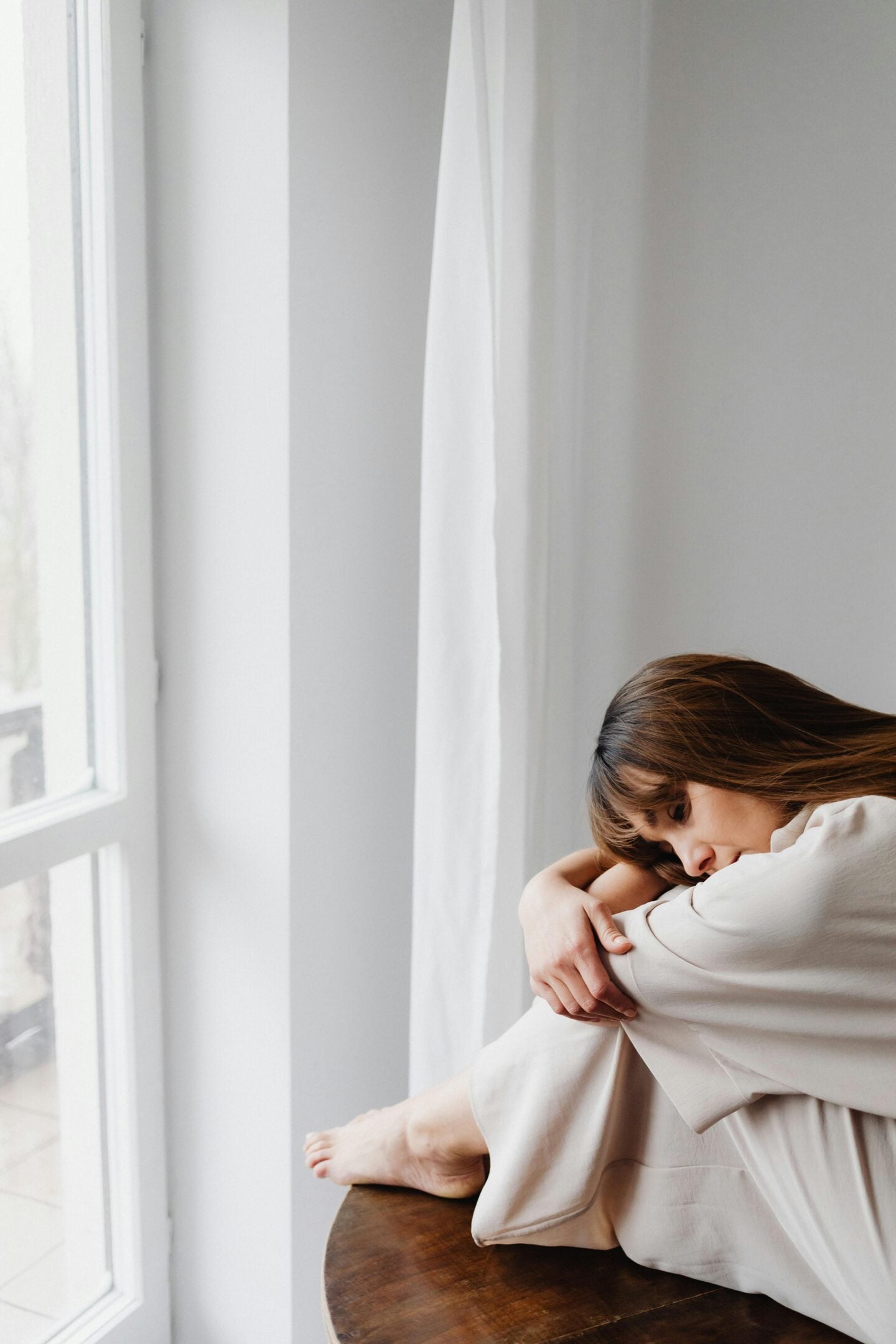Woman sitting alone by a window in a bright minimalist room.