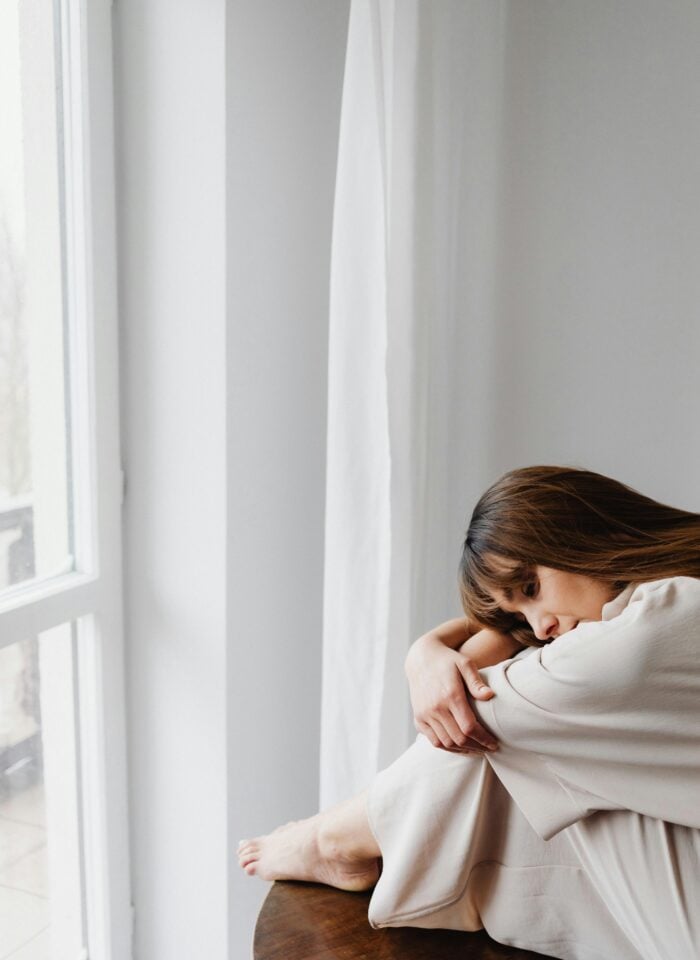 Woman sitting alone by a window in a bright minimalist room.