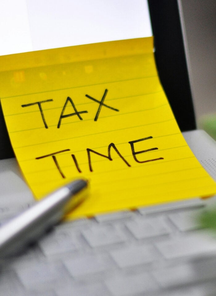 Yellow sticky note with the words “Tax Time” attached to a laptop keyboard beside a pen and blurred green plant.