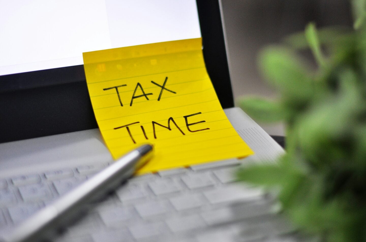 Yellow sticky note with the words “Tax Time” attached to a laptop keyboard beside a pen and blurred green plant.