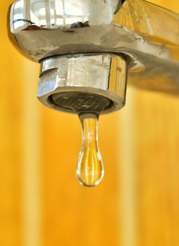 Close-up of a leaking chrome faucet with a single water droplet hanging from the spout against a blurred warm-toned background.