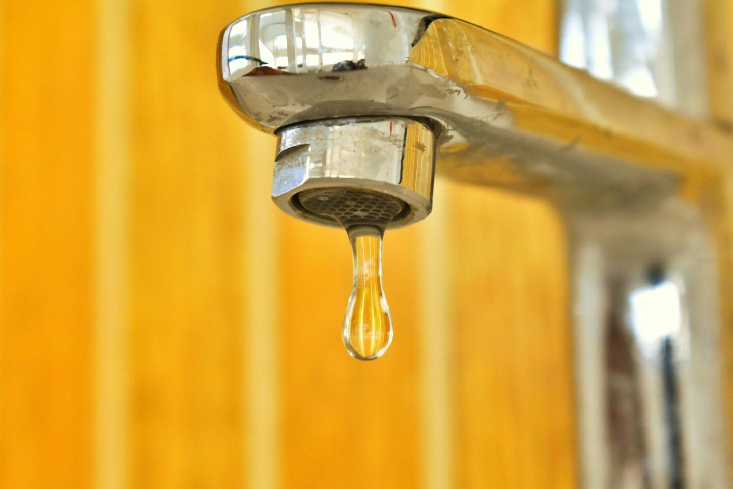 Close-up of a leaking chrome faucet with a single water droplet hanging from the spout against a blurred warm-toned background.