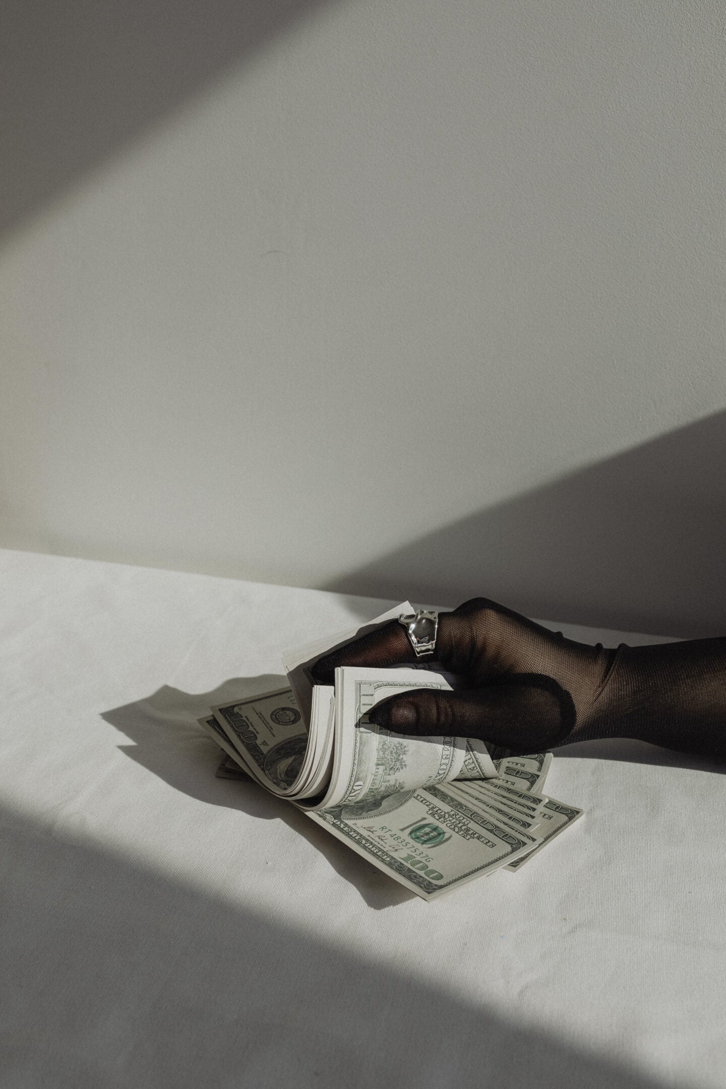 Moody lifestyle photo of a hand in a sheer black glove holding a stack of US dollar bills on a white fabric surface with dramatic natural shadows.