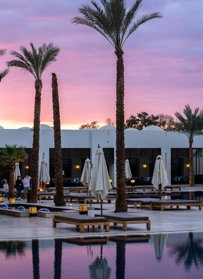 Luxury resort pool area at sunset featuring white modern hotel buildings, tall palm trees, lounge chairs, and calm reflective water under a pink and purple evening sky.