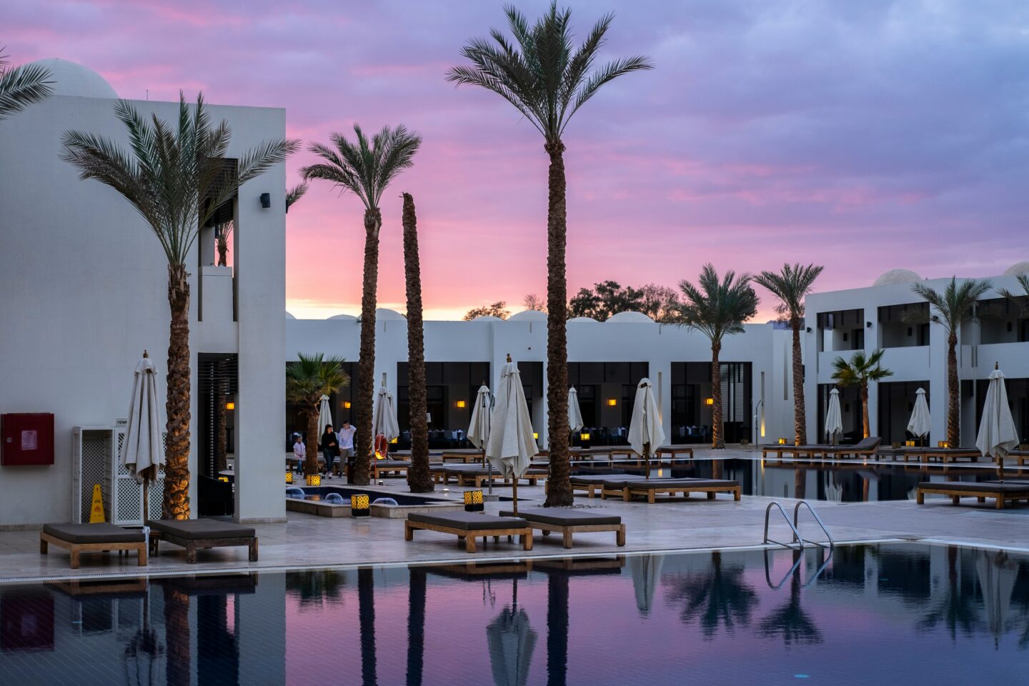 Luxury resort pool area at sunset featuring white modern hotel buildings, tall palm trees, lounge chairs, and calm reflective water under a pink and purple evening sky.