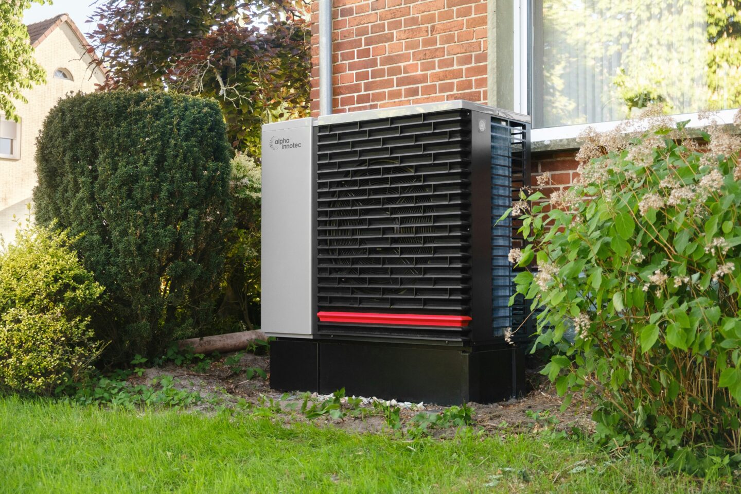 Outdoor residential heat pump unit installed beside a brick house surrounded by garden shrubs and greenery.
