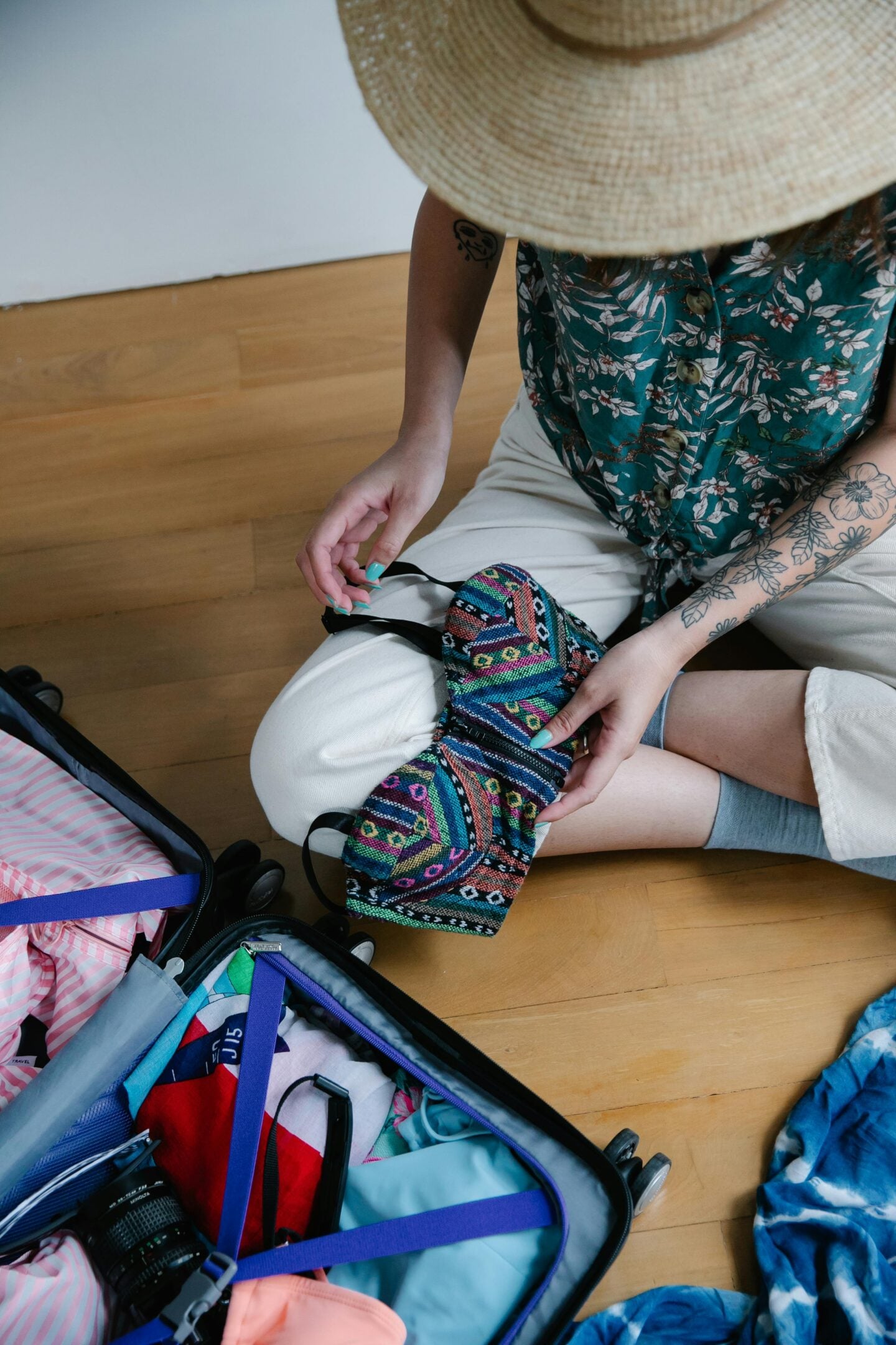 Woman sitting on the floor packing a suitcase for travel while holding a colorful patterned pouch and wearing a straw hat.