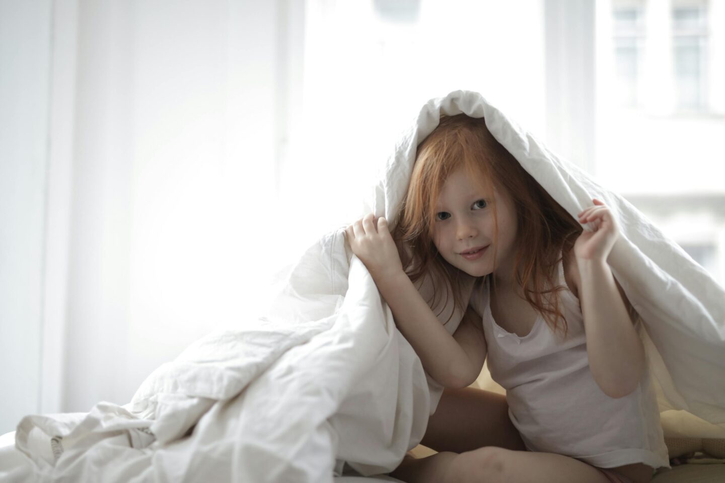 Little red-haired girl sitting under a white duvet on a bed in a bright bedroom, appearing playful and awake during bedtime.