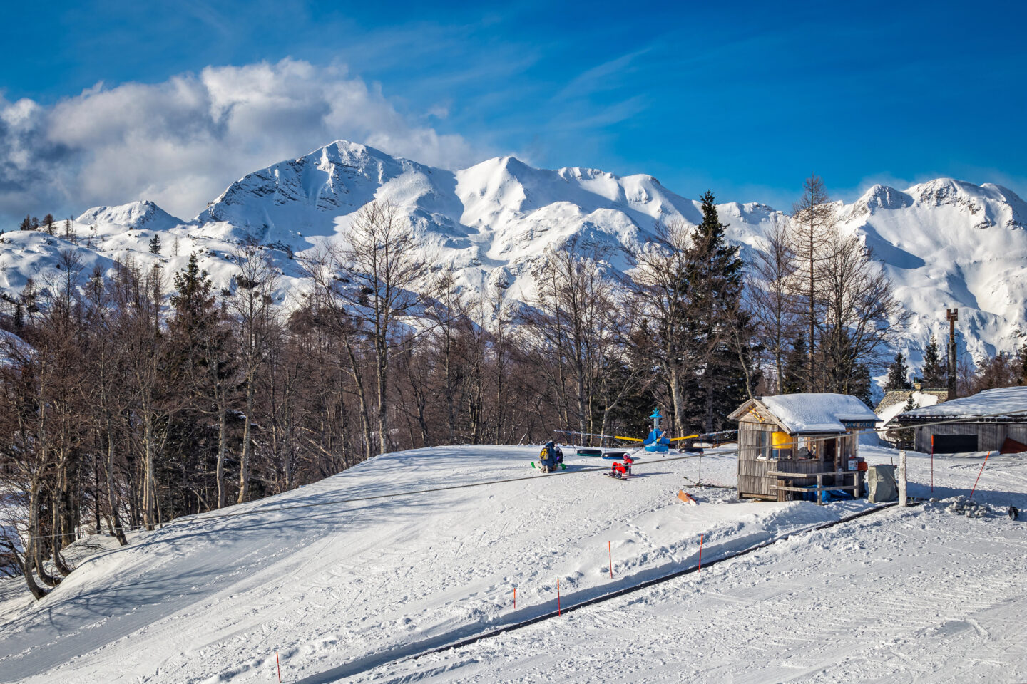 Snow-covered wooden hut at Vogel Ski Resort in Bohinj, Slovenia, surrounded by alpine trees and dramatic mountain peaks under a bright blue winter sky.
