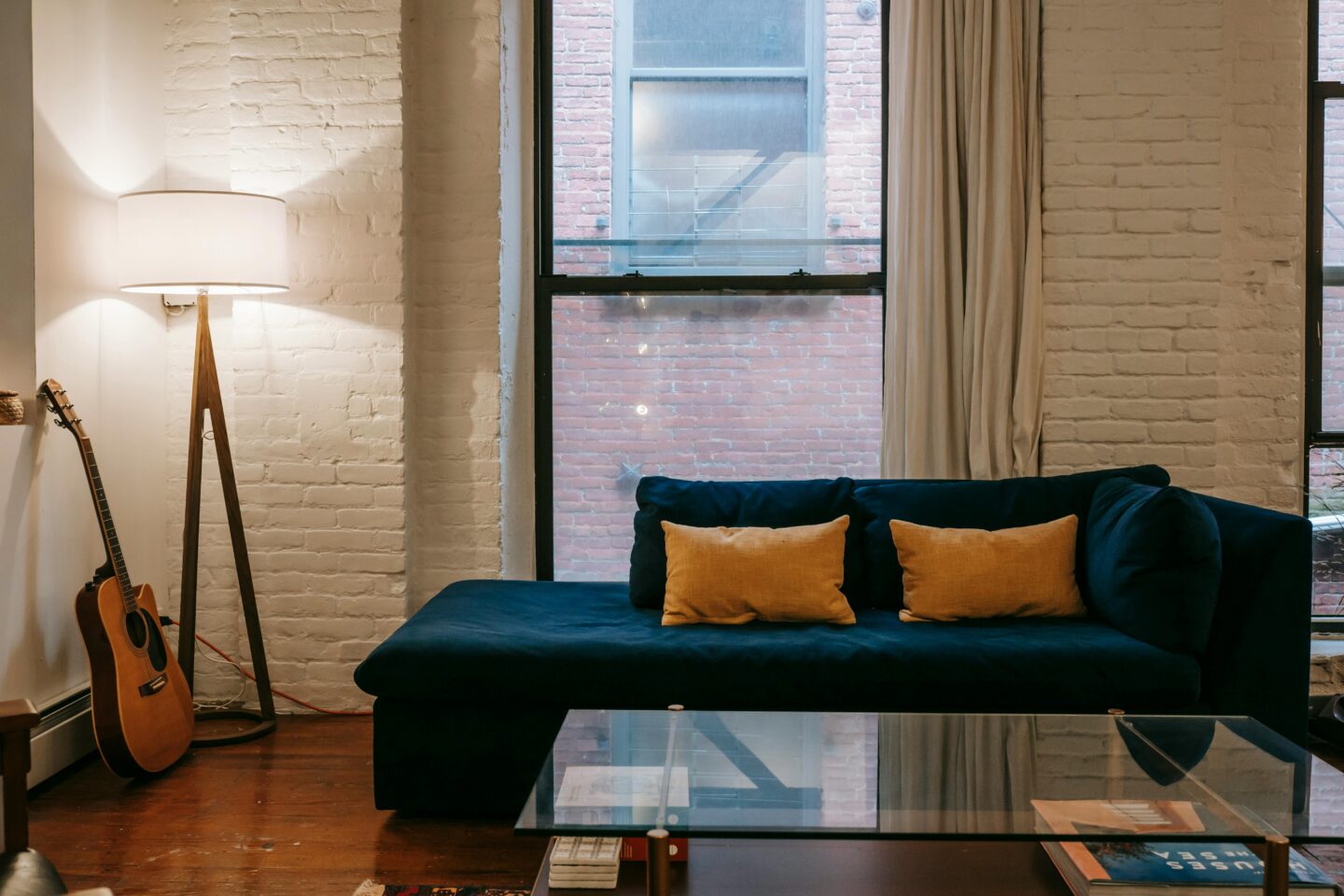 Modern living room with exposed white brick walls, navy sectional sofa, warm floor lamp, and cozy vintage-inspired decor in an older industrial-style apartment.