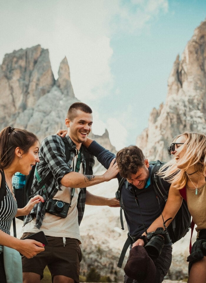 Friends laughing together during a mountain hiking adventure with backpacks and cameras, surrounded by dramatic rocky peaks and scenic alpine views in the background.