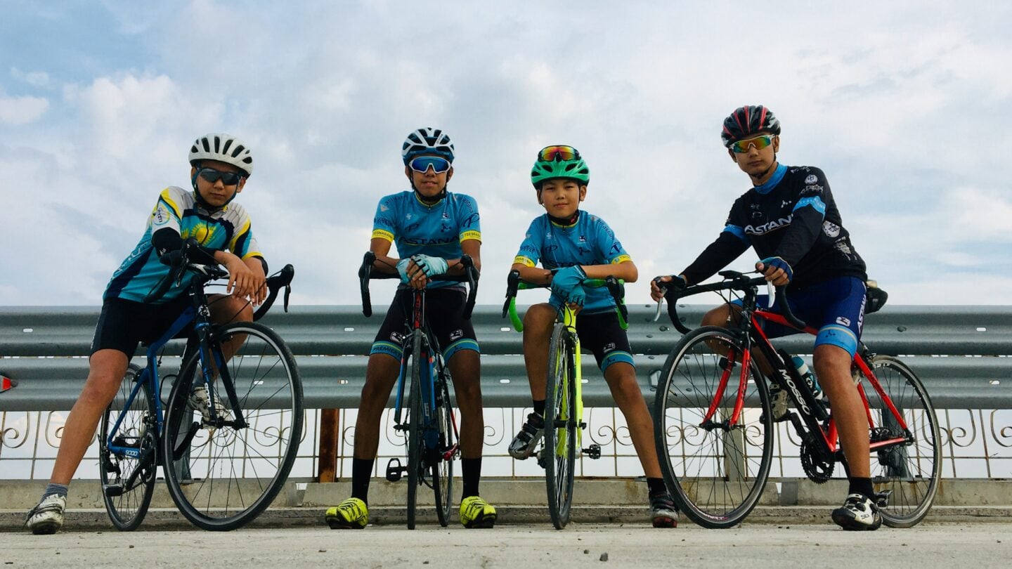 Group of young cyclists posing with road bikes and helmets beside a roadside barrier under a cloudy sky.