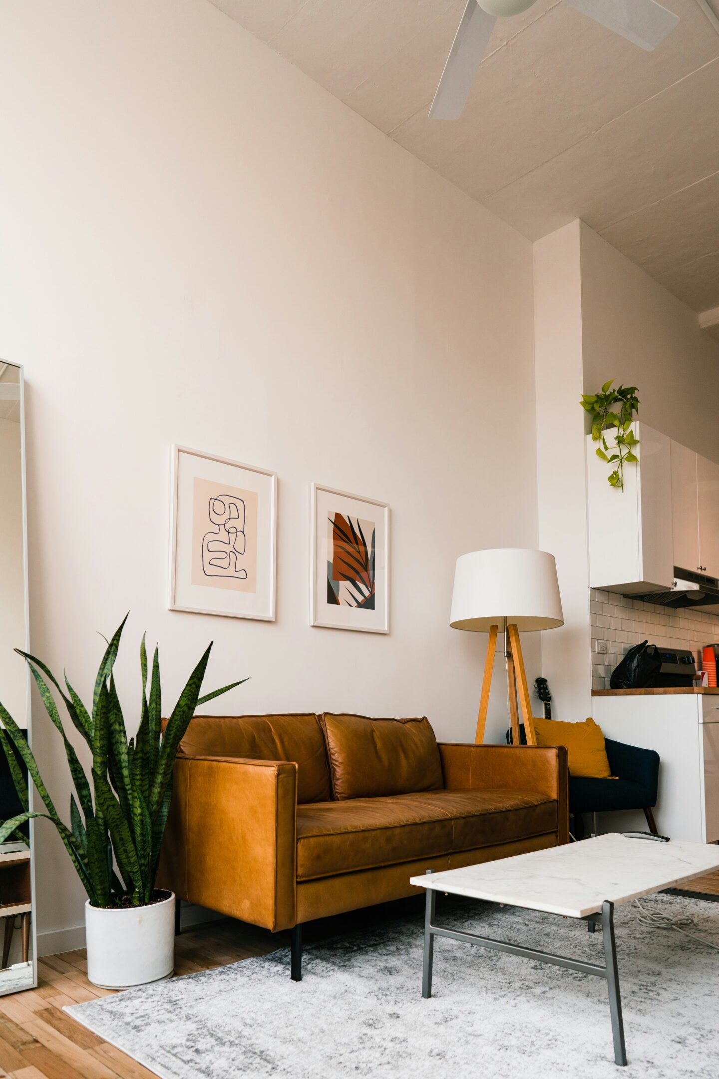 Modern living room with a caramel leather sofa, two minimalist framed artworks on a white wall, a tall snake plant in a white pot, and a marble-top coffee table on a light rug. A wooden tripod floor lamp stands beside the sofa, with a small open kitchen visible in the background featuring white cabinets and a trailing green plant.